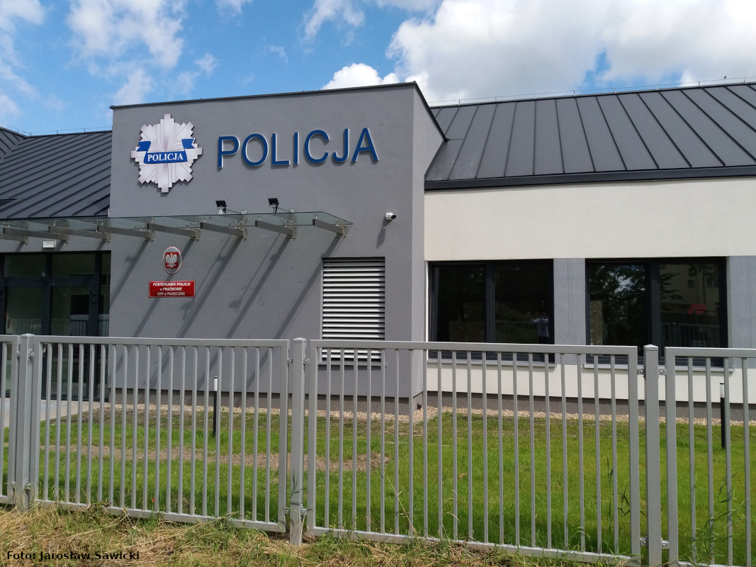 Zdjęcie przedstawia budynek Posterunku Policji w Prażmowie od strony wejścia głównego z większej perspektywy obejmującej drzwi zewnętrzne oraz okna. Na fotografii widać logo Policji, napis „Policja” oraz godło owalne i tablicę urzędu.