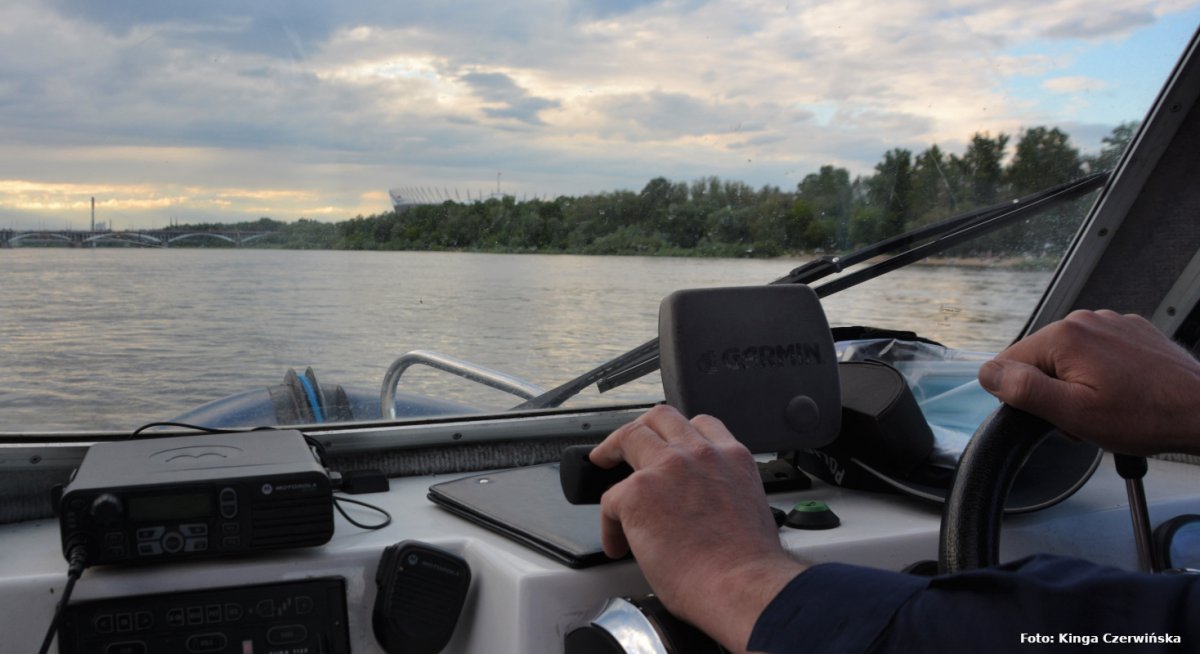 Zdjęcie wykonano z policyjnej łodzi na Wiśle. Na pierwszym palnie widać kokpit i ręce funkcjonariusza, trzymające ster. Na wprost za szybą rozpościera się panorama rzeki Wisły. Z jej  prawego brzegu wyłania się Stadion Narodowy.