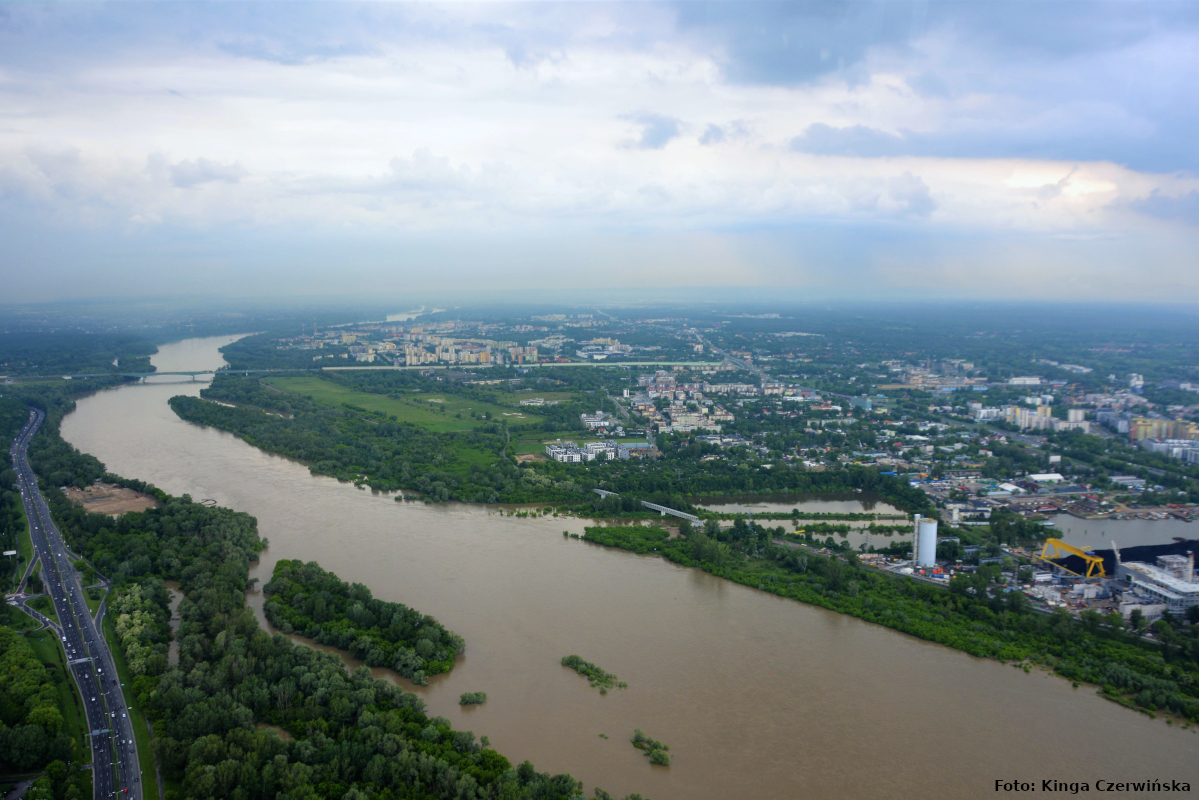 Zdjęcie przedstawia panoramę Warszawy z lotu ptaka. Od prawego dolnego rogu zdjęcia do lewego górnego rozpościera się rzeka Wisła. Po prawej stronie zdjęcia widać liczne zabudowania, po lewej trasę szybkiego ruchu.