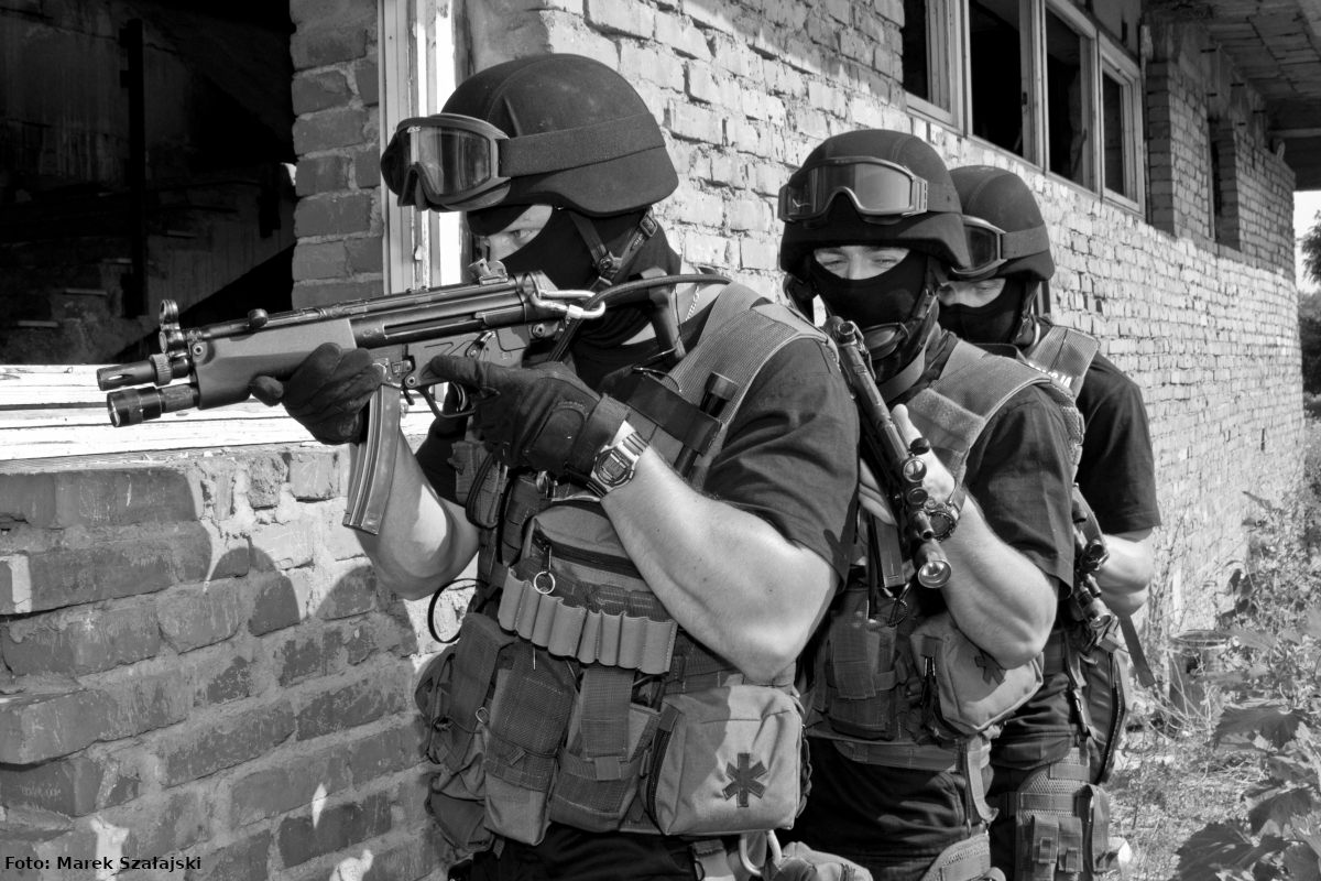 Zdjęcie czarno-białe, wykonane podczas ćwiczeń. Na fotografii widać trzech umundurowanych antyterrorystów z wyposażeniem do służby. Policjanci stoją jeden za drugim przed budynkiem gotowi do pojęcia interwencji.