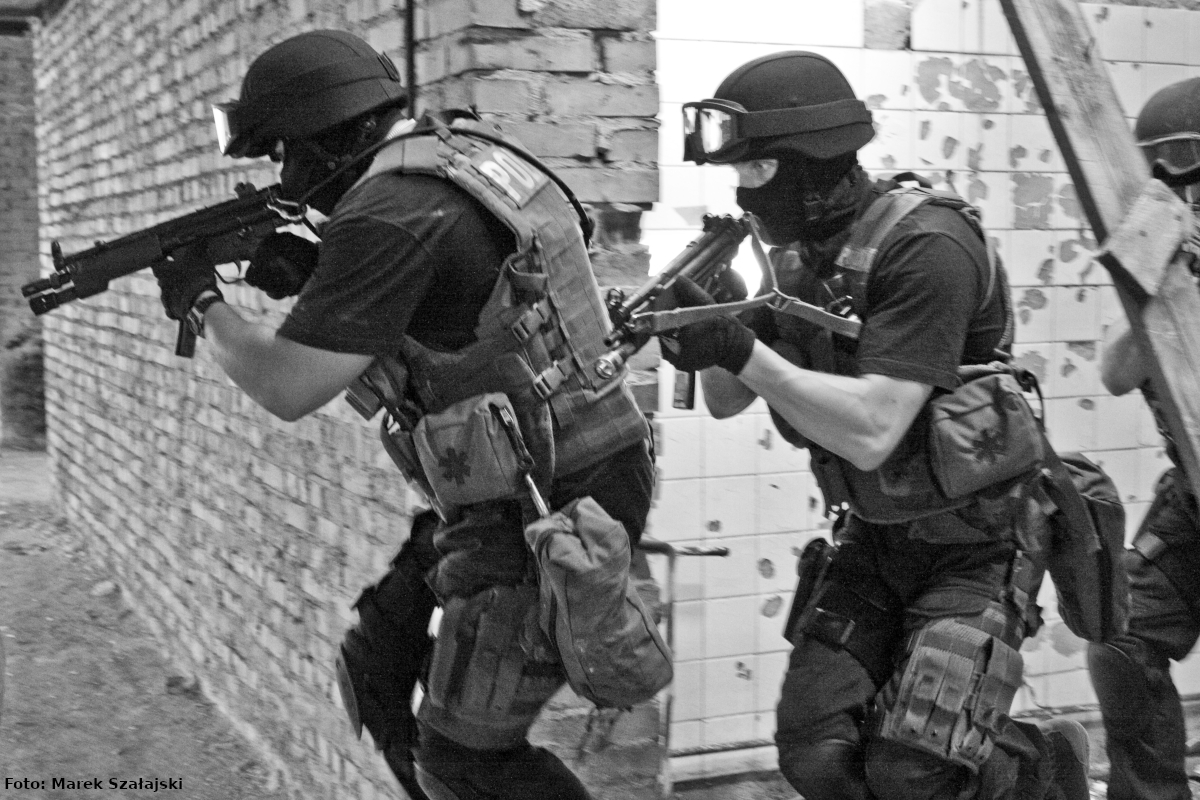 Zdjęcie czarno-białe wykonane przed budynkiem podczas ćwiczeń. Przedstawia antyterrorystów z wyposażeniem do służby, którzy biegną, jeden za drugim, z bronią gotową do ewentualnego użycia. 