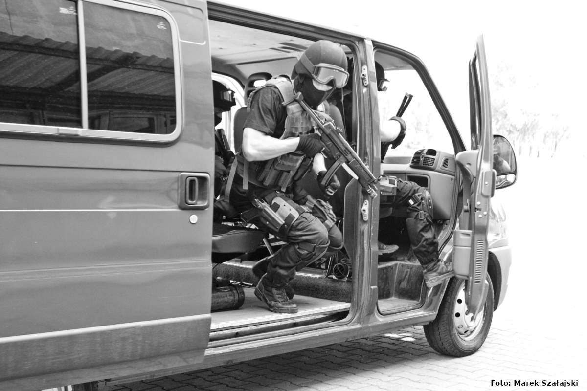 Zdjęcie czarno-białe, z radiowozu nieoznakowanego typu van wybiegają umundurowani antyterroryści z wyposażeniem do służby gotowi do podjęcia interwencji.