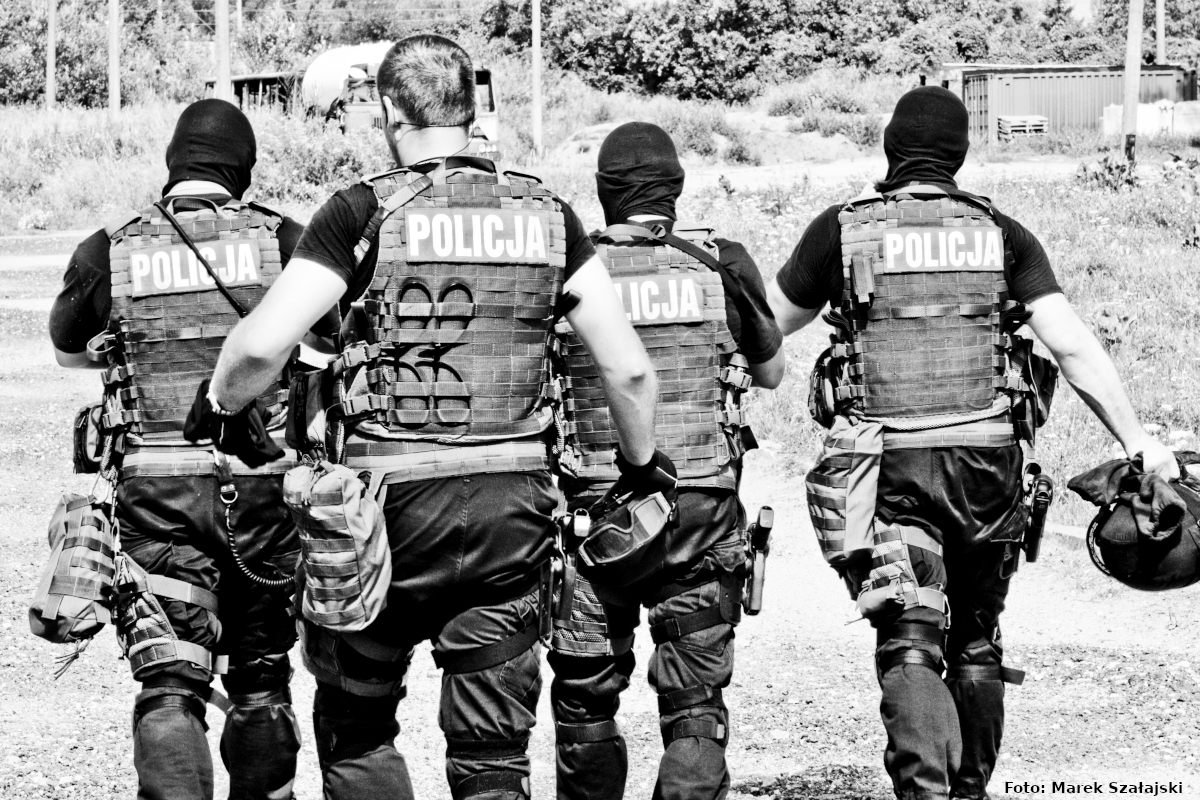 Zdjęcie czarno-białe przedstawia 4 antyterrorystów odwróconych plecami. Policjanci zdejmują hełmy i wracają do jednostki po zakończonej akcji. 
