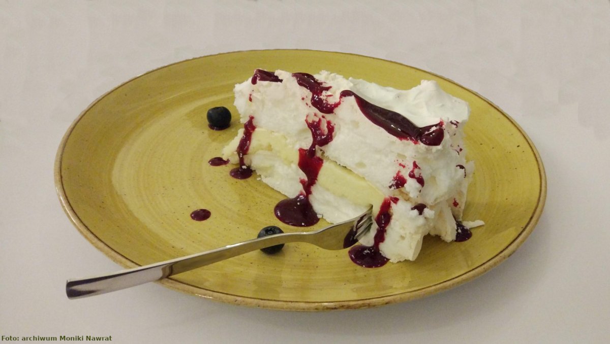 Na zdjęciu widać talerzyk deserowy koloru żółtego, na którym znajduje się deser lodowy z polewą jagodową, obok leżą dwie jagody. Na talerzu położony jest widelczyk częściowo dotykający deseru.