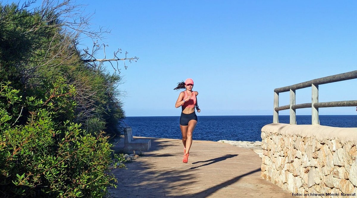 Na zdjęciu widać kobietę w sportowym stroju, która biegnie między krzewami i murkiem przy nadmorskim deptaku, z tyłu widać morze