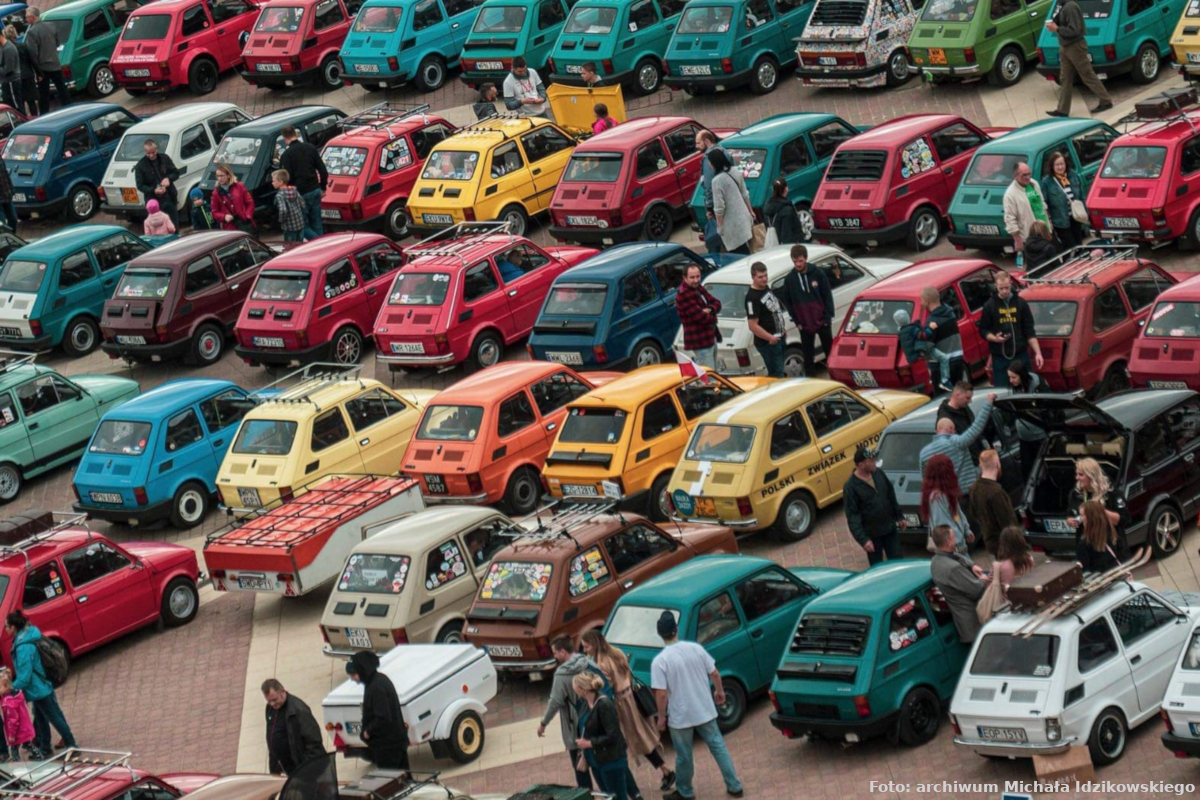 Zdjęcie wykonano podczas „Zlotu Fiata 126P”, widać na nim dużo małych fiatów w różnych kolorach. Samochody zaparkowane są obok siebie w kilku rzędach. 