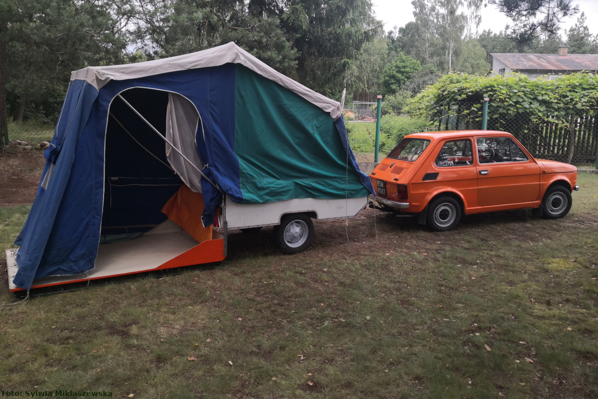 Zdjęcie wykonano w plenerze. Widać na nim pomarańczowego Fiata 126P z rozłożoną przyczepą kempingową Niewiadów N126 w kolorze niebiesko-biało-zielonym z pomarańczowymi elementami.