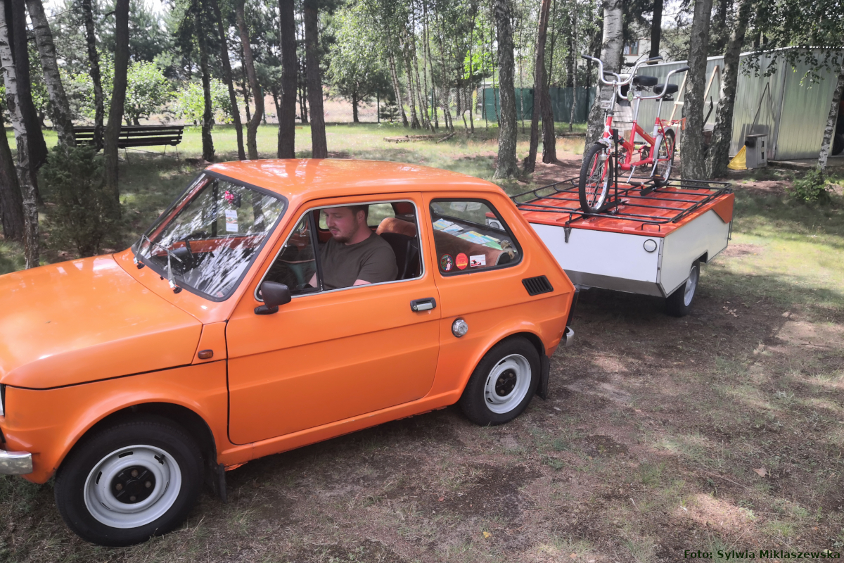 Na zdjęciu widać pomarańczowego Fiata 126P z podpiętą przyczepą kempingową Niewiadów N126 z zamontowanym rowerem. Za kierownica siedzi Michał.