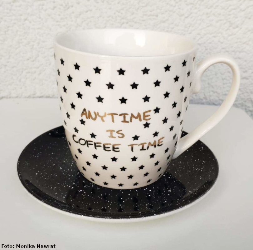 Na zdjęciu widać na czarnym podstawku kubek biały z nadrukowanymi czarnymi gwiazdkami z napisem „anytime is coffee time” ( ang. każdy czas jest dobry na kawę).
