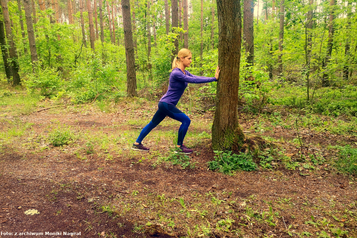 Zdjęcie obrazuje rozciąganie mięśni goleni  Monika opiera ramiona o drzewo i napiera na nie, prawą nogę ma ugiętą, a lewą wyprostowaną na całej stopie i napiera na drzewo.