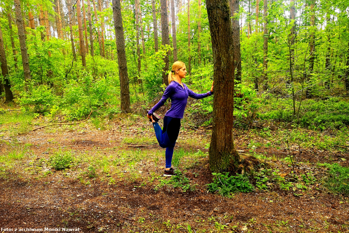 Zdjęcie obrazuje rozciąganie mięśnia czworogłowego uda Monika stoi w pozycji wyprostowanej, dociąga piętę do pośladka, kolana trzyma blisko siebie. Dla utrzymania równowagi podtrzymuje się drzewa.