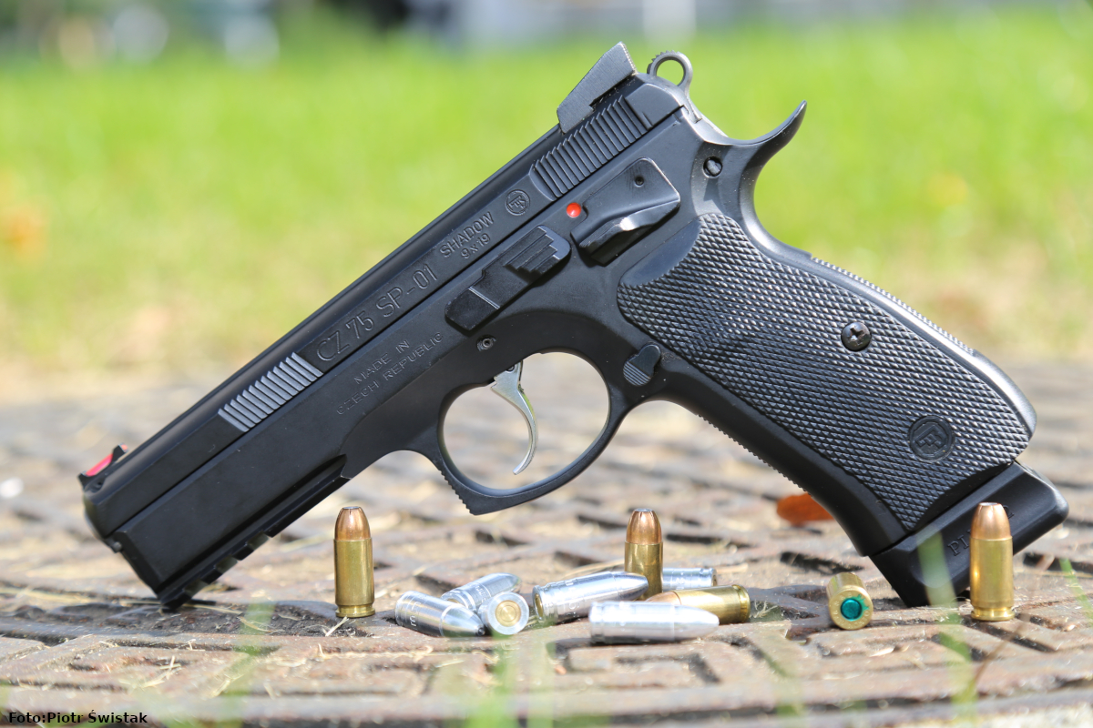 Na zdjęciu na pierwszym planie widać pistole model CZ 75 SP 01 Shadow, koloru czarnego oparty lufą i częścią magazynka na stalowej płycie. Drugi plan jest rozmazany, przedstawia trawę.