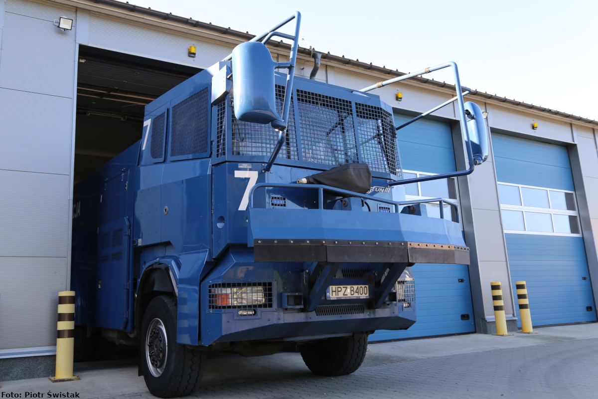 Zdjęcie przedstawia przód opancerzonego samochodu ciężarowego marki Scania tzw. „Tajfun III” koloru niebieskiego, który wyjeżdża z garażu. Pojazd ten jest wyposażony m.in.: w armatki wodne, kamery telewizji przemysłowej, taran.