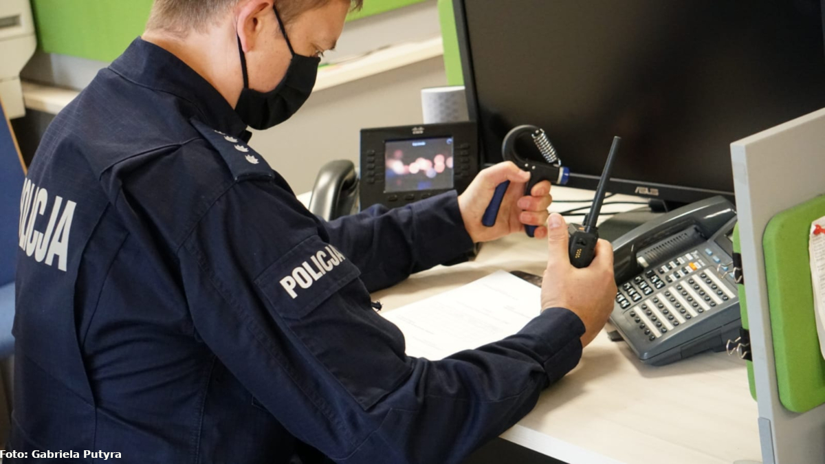 Na zdjęciu widać umundurowanego policjanta, który siedzi przy biurku w prawej ręce trzyma radiostację, a w lewej ręce gripmaster. 