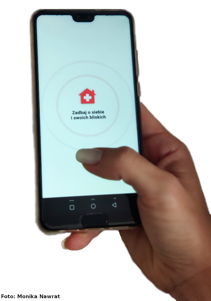 Na zdjęciu widać kobiecą dłoń z telefonem komórkowym. Na ekranie smartfona jest wyświetlana aplikacja z komunikatem „zadbaj o siebie i swoich bliskich” oraz logiem w postaci szkicu domku jednorodzinnego w kolorze czerwonym z białym krzyżem pośrodku. 