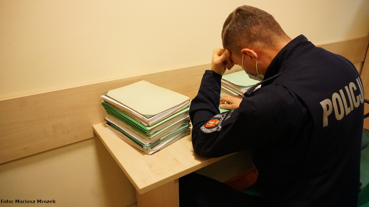Na zdjęciu  widać umundurowanego policjanta, który siedzi przy biurku, na którym leżą akta, i przegląda dokumenty z głowa opartą na lewej dłoni.