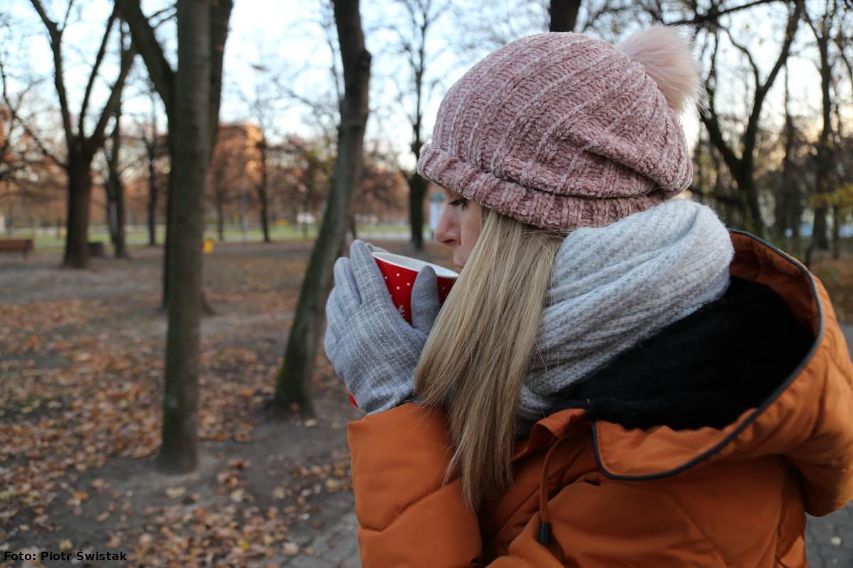 Zdjęcie przedstawia kobietę ubraną w czapkę, szalik, rękawiczki i kurtkę zimową, która pije ciepły napój w kubku koloru czerwonego w białe kropki. Na drugim planie widać drzewa. 