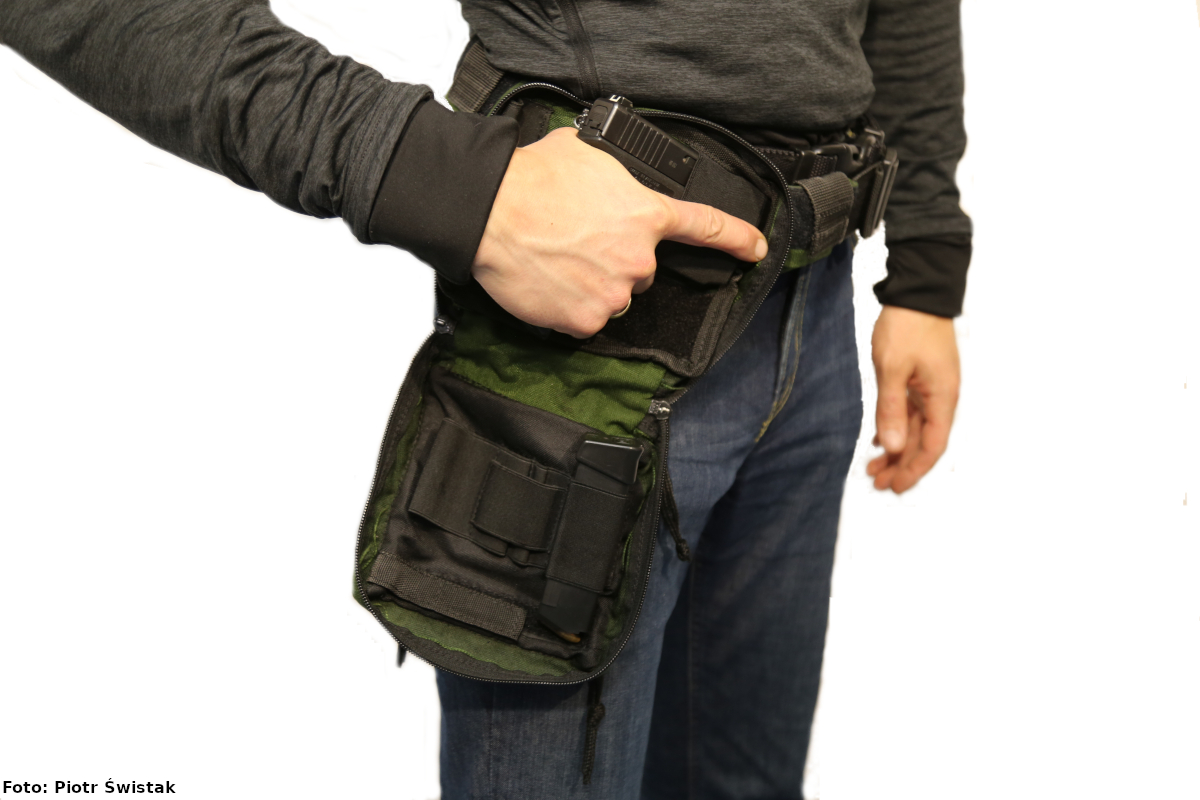 Zdjęcie przedstawia nieumundurowanego policjanta, który trzyma w prawej dłoni broń przypiętą do zielono-czarnej kabury mocowanej na pasku.